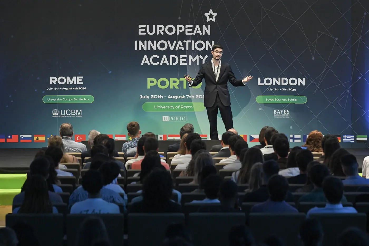 A speaker on stage in front of a large European Innovation Academy screen