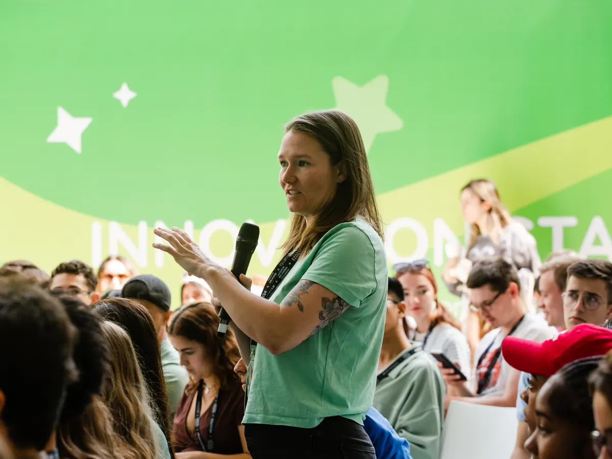 A participant with a microphone asks a question from a packed audience